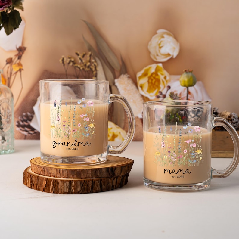 May include: Two clear glass mugs filled with a light brown liquid, each with a handle. One mug says "grandma" and the other says "mama" in black script, with floral designs. The mugs sit on a wooden coaster.