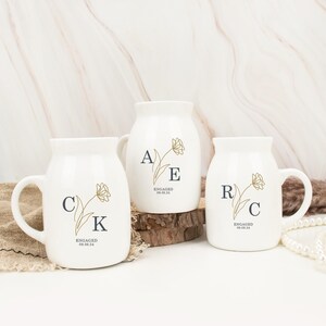 May include: Three white ceramic mugs with gold floral designs and black lettering. The mugs say "A E ENGAGED 08.08.24", "C K ENGAGED 08.08.24", and "R C ENGAGED 08.08.24".