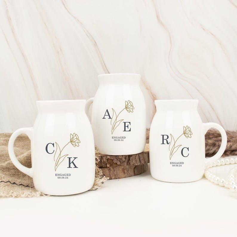 May include: Three white ceramic mugs with gold floral designs and black lettering. The mugs say "A E ENGAGED 08.08.24", "C K ENGAGED 08.08.24", and "R C ENGAGED 08.08.24".