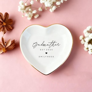 Puede incluir: Un plato de cerámica blanco en forma de corazón con borde dorado. El plato está inscrito con la palabra "Godmother", el año "EST 2025", un símbolo de corazón y el nombre "EMILY ROSE" en escritura negra. El fondo es rosa con flores secas y flores blancas.
