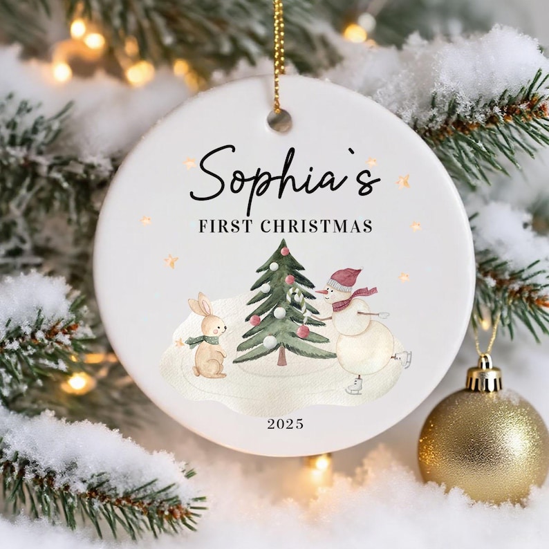 May include: A white ceramic Christmas ornament with the text "Sophia's FIRST CHRISTMAS" and the year 2025. The ornament features a watercolor illustration of a snowman, a rabbit, and a Christmas tree. The ornament is hanging from a gold chain.