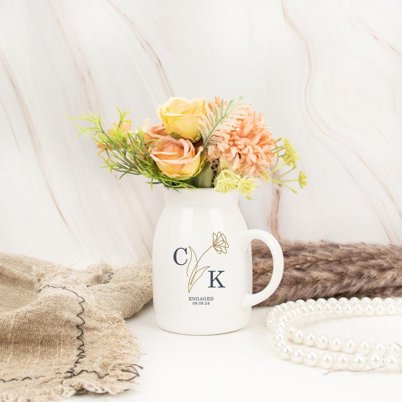 May include: White ceramic vase with a gold floral design and the initials "C K" and the text "ENGAGED 08.04.24". The vase is filled with a bouquet of peach and yellow flowers.