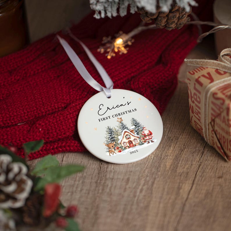 May include: A round white ceramic Christmas ornament hanging by a white ribbon. The ornament features the text "Erica's FIRST CHRISTMAS" above a colorful illustration of a snowy cabin, pine trees, a reindeer, and Santa Claus, with "2025" below.