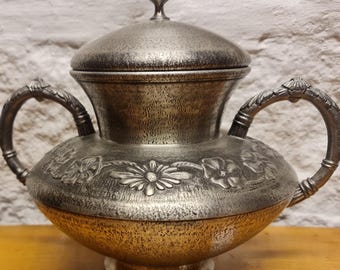 May include: A silver sugar bowl with a lid and two handles. The bowl is decorated with embossed floral designs.