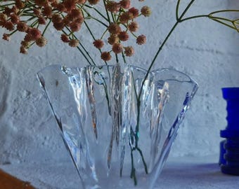 Op de afbeelding: Een heldere glazen vaas met een gegolfde rand bevat een boeket van delicate roze bloemen. De vaas staat op een wit oppervlak met drie blauwe glazen vazen op de achtergrond.
