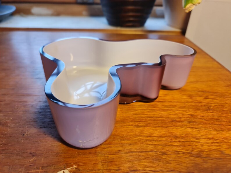 May include: A white and purple glass bowl with an abstract, organic shape. The bowl is made of two pieces of glass, one white and one purple, that are joined together.