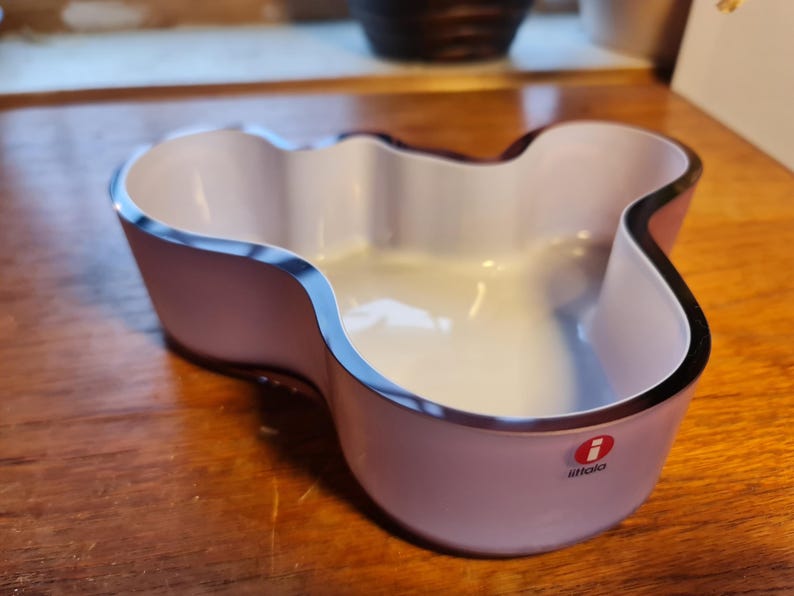 May include: A white and purple glass bowl with a wavy, organic shape. The bowl has a black rim and the iittala logo on the bottom.