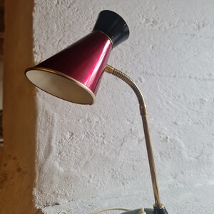 Puede incluir: Una lámpara de escritorio vintage roja y negra con un brazo flexible y un interior blanco. La lámpara tiene una base negra y un tallo dorado.