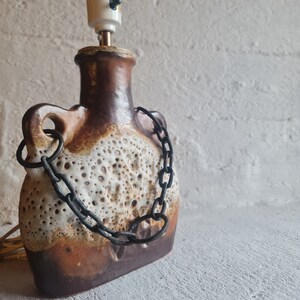 Puede incluir: Una lámpara de mesa de cerámica marrón y blanca con un asa de cadena. La lámpara tiene una superficie texturizada y una pantalla de cerámica blanca.