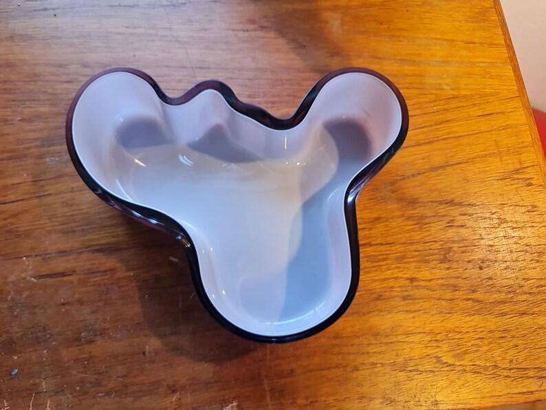 May include: A purple glass bowl with a unique, organic shape. The bowl has a smooth, white interior and a dark purple rim.