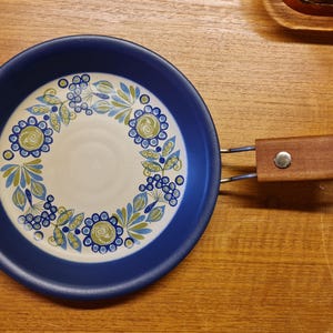 May include: A blue and white ceramic pan with a floral design. The pan has a wooden handle attached with metal brackets. The floral design includes blue and green flowers and leaves. The pan is on a wooden surface.