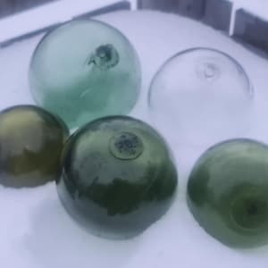 May include: Five glass fishing floats in various shades of green. Some floats have a faded, aged look. One float has a small, faded label with the text "P.O.S."