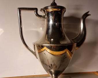 Op de afbeelding: Een zilveren theepot met een rond lichaam en een decoratief handvat. De theepot heeft een glanzende afwerking en een bloemenmotief aan de zijkant.