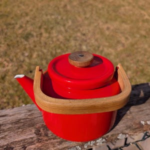 Puede incluir: Una tetera roja vibrante con asa y pomo de madera. La tetera tiene un pico blanco y se asienta sobre una superficie de madera. El asa es de madera clara, contrastando con el acabado esmaltado rojo.