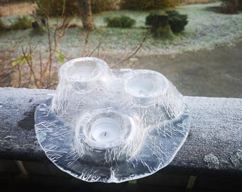 May include: A clear, glass candle holder with three candle wells. The holder has a textured, icy appearance and sits on a round base. The candles are white. The holder is approximately 8 inches in diameter.