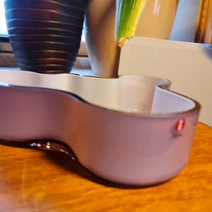 May include: A light pink glass bowl with a unique, wavy design. The bowl is made by Iittala and has a white interior.