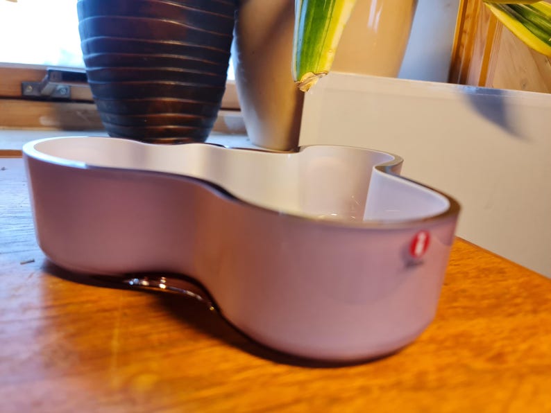 May include: A light pink glass bowl with a unique, wavy design. The bowl is made by Iittala and has a white interior.