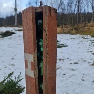 Può includere: Una scatola di legno con una lettera H bianca dipinta sul lato. La scatola è aperta e contiene diverse bottiglie di vetro verde impilate all'interno.