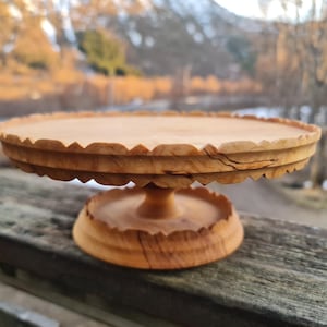 Könnte beinhalten: Ein Kuchenständer aus Holz mit einem gewellten Rand und einem Sockel. Der Ständer ist aus hellbraunem Holz mit einer natürlichen Oberfläche.