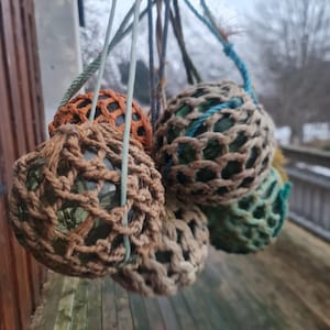 Op de afbeelding: Een groep van zes glazen boeien, elk omhuld in een gehäkeld net van bruin, beige en blauw, hangend aan een houten oppervlak.
