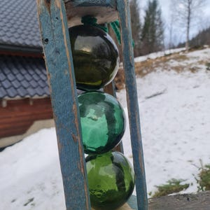 Op de afbeelding: Drie groene glazen boeien zijn gestapeld in een verweerd houten frame. Het frame is blauw geverfd en heeft een versleten, rustieke uitstraling. De boeien zijn in verschillende tinten groen en hebben een licht troebele uitstraling.