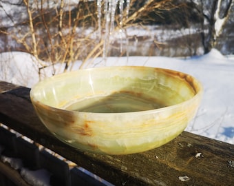 May include: A large, round, green and brown onyx bowl with a smooth, polished surface. The bowl is filled with water and is sitting on a wooden surface.