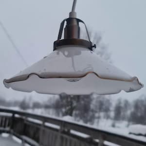 Op de afbeelding: Een witte lampenkap met gegolfde rand en een koperkleurige metalen fitting hangt aan een wit snoer. De lampenkap is nat, wat suggereert dat hij buiten in een besneeuwde omgeving hangt.