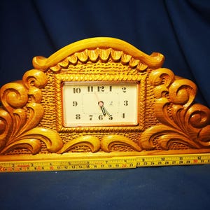 May include: A wooden mantel clock with a light brown finish. The clock has an ornate, carved design with a rectangular clock face. The clock face has black numerals and hands. A ruler is placed in front of the clock.