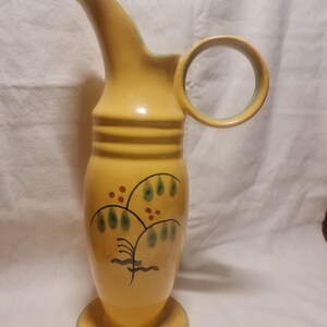 May include: A yellow ceramic pitcher with a green handle and a painted design of a tree with green leaves and red berries. The pitcher has a wide base and a narrow neck.