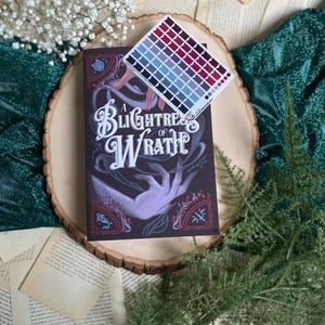 May include: A book titled "A Blight of Wrath" with a dark blue cover and a hand reaching out from the book. The book is on a wooden surface with a color palette card on top of it.