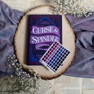 May include: A purple and blue book titled "Curse & Spindle: A Realm of Revelry" by Chelsea  Cain. The book is on a wooden surface with a small color palette sticker.