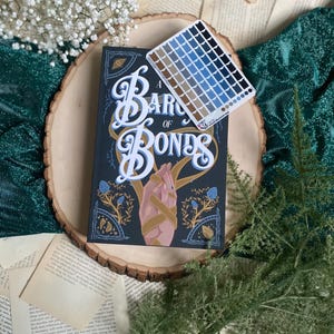 May include: A hardcover book titled "A Barony of Bones" with a dark blue cover and gold lettering. The book is sitting on a wooden slice with a color palette card next to it.