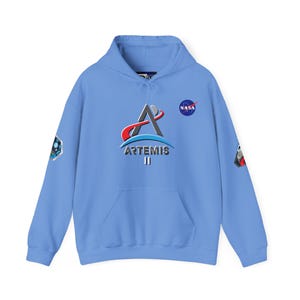 Pode incluir: Um moletom azul claro com o logotipo Artemis II em vermelho, azul e prata e o logotipo da NASA. O moletom tem patches nas mangas, um bolso frontal e um capuz com cordão.