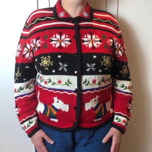 Könnte beinhalten: Eine rote, schwarze und weiße Weihnachtskardigan mit Schneeflocken- und Hundemustern. Der Strickpullover hat einen Kragen und lange Ärmel. Das Design umfasst gestickte Schneeflocken, florale Akzente und zwei weiße Hunde.