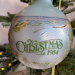 Könnte beinhalten: Hellblaue Weihnachtskugel mit silberner Kappe. Sie zeigt ein florales Design in Grün, Blau und Lila, mit dem Wort "CHRISTMAS" und dem Jahr "1984" in grüner Schrift.