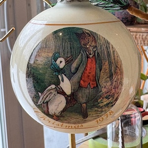 Puede incluir: Adorno navideño color crema con una ilustración detallada de un zorro y un pato en una escena de bosque. El adorno tiene letras doradas que dicen "Christmas 1978". El adorno está colgado de un soporte dorado.