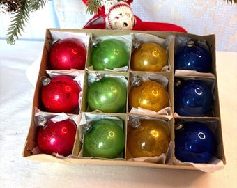 Pode incluir: Uma caixa de cartão cheia de doze enfeites de Natal vintage em vermelho, verde, dourado e azul. Os enfeites são redondos e brilhantes, com tampas de metal prateado. Uma pequena árvore de Natal e uma figura decorativa estão no fundo.