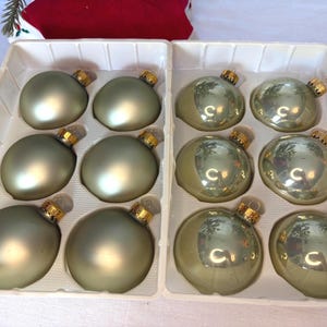 May include: A set of twelve round Christmas baubles arranged in a white plastic tray. The baubles are divided into two groups: six have a matte sage green finish, and six have a reflective, glossy metallic sage green finish with gold caps.