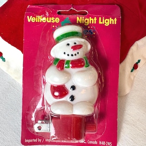 May include: A snowman night light in a red and pink package. The snowman has a green and white hat, a red scarf, and black button eyes. The package reads "Night Light" and "Veilleuse".