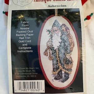 Op de afbeelding: Een "Antique Santa Kit" van Cross My Heart, met een kruissteekontwerp van de Kerstman. De kit bevat stof, floss, een naald en andere materialen. De verpakking heeft een rode rand en een gouden koord. De kit is uit 1993.