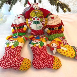 Peut inclure: Une collection de décorations de Noël faites à la main, comprenant des figurines du Père Noël et d'anges, ainsi que des bas de Noël. Les ornements sont en tissu avec des motifs rouges, blancs, jaunes et verts. Les figurines du Père Noël ont des ceintures noires et des costumes rouges.