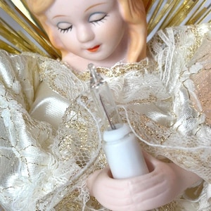 Könnte beinhalten: Ein Vintage-Weihnachtsbaumspitze in Form eines Engels. Der Engel hat ein Porzellangesicht mit geschlossenen Augen und einem ruhigen Ausdruck. Sie trägt ein weißes und goldenes Spitzenkleid und hält eine kleine Glühbirne. Goldener Lametta bildet die Flügel des Engels.