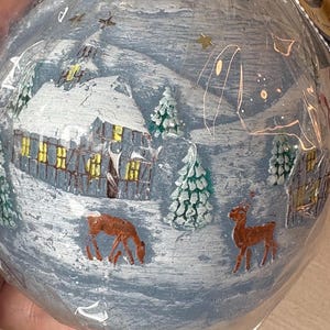 May include: A round, hand-painted Christmas ornament with a winter scene. The ornament features a snow-covered village with a house, trees, and deer. The color palette includes blue, white, brown, and gold accents. The ornament is wrapped in clear plastic.