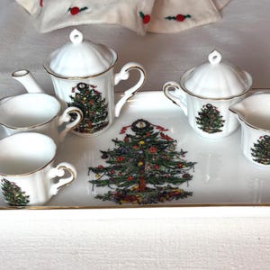 May include: A white porcelain tea set comprising a rectangular tray, a teapot with a lid, a lidded sugar bowl, a creamer, and two cups. Each piece is decorated with a colourful Christmas tree graphic and finished with a gold-toned rim.
