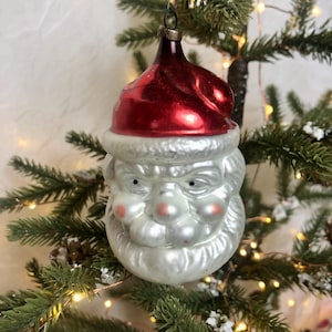 May include: A vintage Christmas bauble in the shape of Santa Claus's head. The bauble features a red hat, a white beard, and pink cheeks. It is suspended from a Christmas tree with green needles and fairy lights.