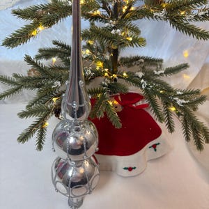 Puede incluir: Un adorno plateado para la parte superior del árbol de Navidad con una parte superior larga y puntiaguda y tres secciones esféricas. El adorno se coloca en un pequeño árbol de Navidad con agujas verdes, luces y una falda roja.