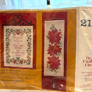 Pode incluir: Um kit de ponto cruz embalado com dois desenhos. Um desenho tem o texto "May your Christmas be filled with Wonder and touched with Love". O outro desenho apresenta poinsétias vermelhas. A embalagem tem o número 2163.