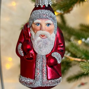 May include: A festive red Santa Claus Christmas bauble with silver glitter detailing. The ornament depicts a detailed face with blue eyes and a white beard. The Santa figure is wearing a red coat and hat, and is holding a red sack.