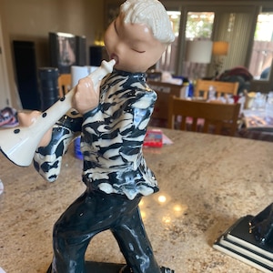 May include: A ceramic figurine of a person playing a white trumpet. The figurine is wearing a black and white patterned shirt and black pants. The figurine is standing on a black and white base.
