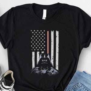 Puede incluir: Camiseta negra con un dise&ntilde;o gr&aacute;fico del casco de Darth Vader integrado en una bandera estadounidense. Las rayas de la bandera son blancas y el sable de luz es rojo. La camiseta se muestra con zapatillas negras y vaqueros azules.